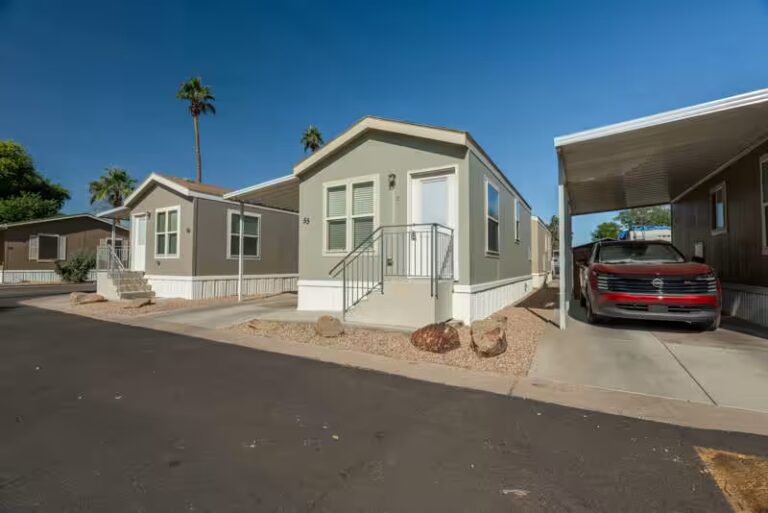 Gray mobile home with red SUV under carport, palm trees, and desert landscaping under blue sky.