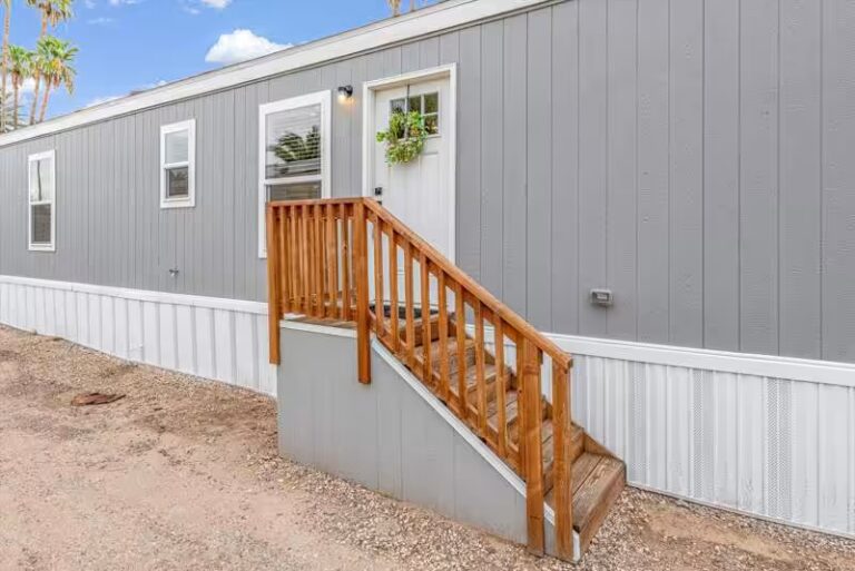 Gray mobile home with wooden stairs and railing leading to white front door, palm trees behind