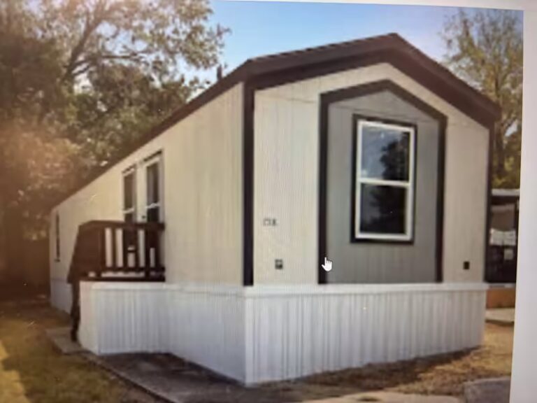 White mobile home with black trim, gray gable window, and wooden deck stairs surrounded by trees.