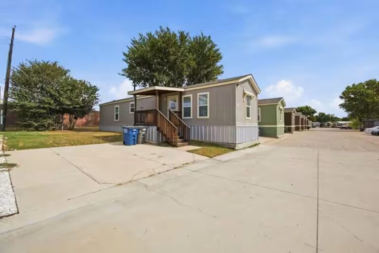 White single-wide mobile home behind chain-link fence at a quiet residential street intersection under blue sky.