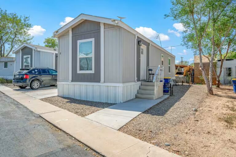 Small mobile home with light siding, gray shutters, front porch ramp, parked truck nearby under clear sky.