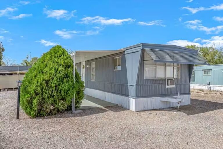 Mobile home behind large trimmed evergreen shrub on gravel lot beneath partly cloudy blue sky.