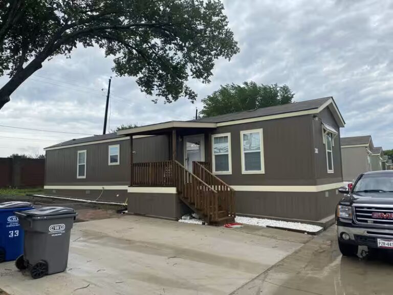 Single-wide trailer home with dark gray siding sits between parked cars beneath an overcast sky.