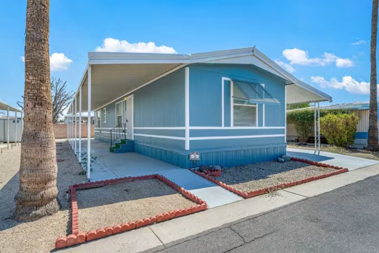 Light blue manufactured home with white accents sits beneath a clear sunny sky in quiet park setting