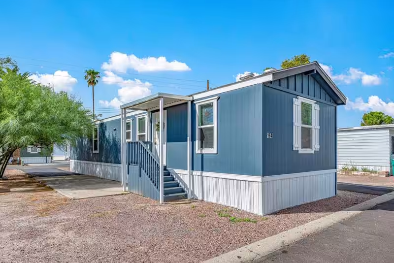 Blue single-wide mobile home with white skirting sits beneath a bright blue sky near desert trees.