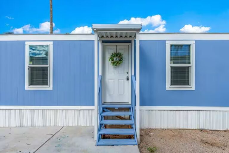 Light blue mobile home with white front door, green wreath, and wooden stairs leading to entrance.