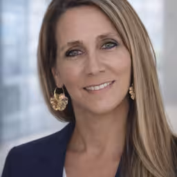 Smiling professional woman with long brown hair wearing a dark blazer and gold earrings in headshot portrait.
