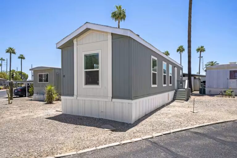 Gray mobile home with white trim, entry stairs, and palm trees in a sunny desert community.