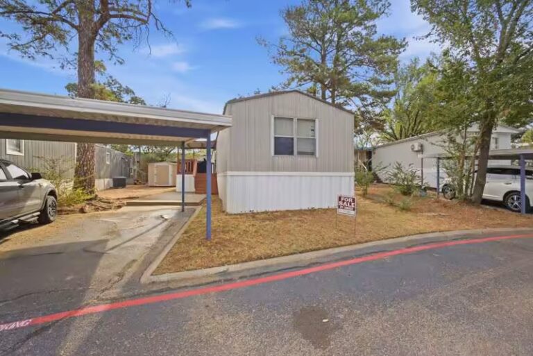 Small beige mobile home with FOR SALE sign in yard, next to carport and trees.