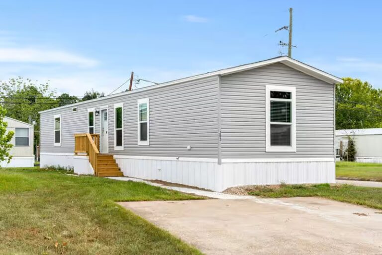 Gray single-wide mobile home with wooden stairs, white trim, and driveway under blue sky.