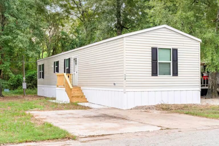 Single-wide mobile home with beige siding, black shutters, and wooden stairs surrounded by trees.