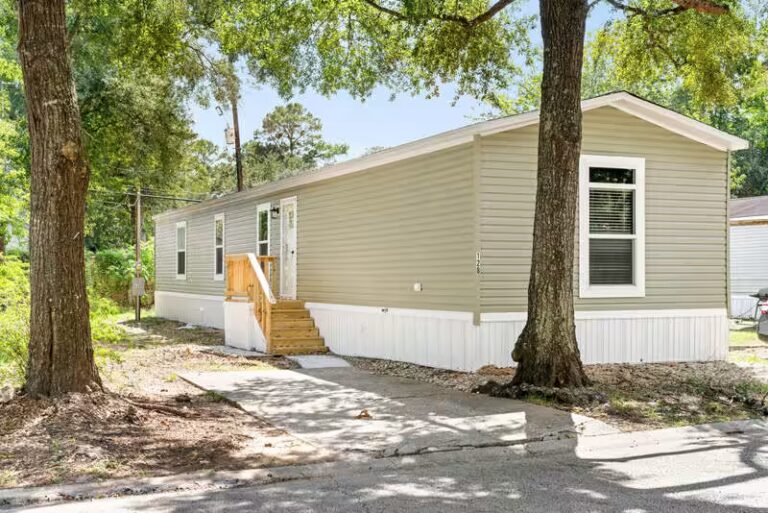 Beige single-wide mobile home with white skirting and wooden steps, nestled among tall trees.