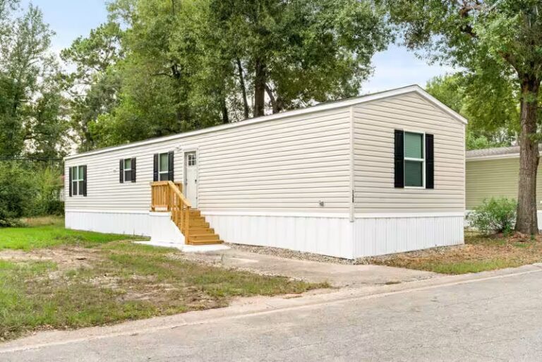 Single-wide mobile home with beige siding, black shutters, and wooden stairs, surrounded by trees.
