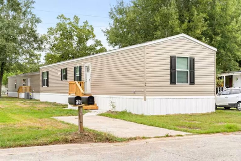 Tan mobile home with black shutters, wooden deck, and mailbox numbered 168 beside tree-lined street.