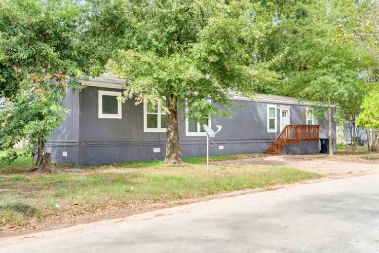Gray mobile home with wooden deck stairs surrounded by trees, satellite dish in front yard.