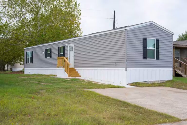Gray mobile home with black shutters and wooden porch steps on a grassy lot.