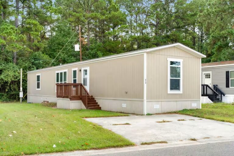 Beige mobile home with wooden deck and stairs surrounded by tall pine trees.