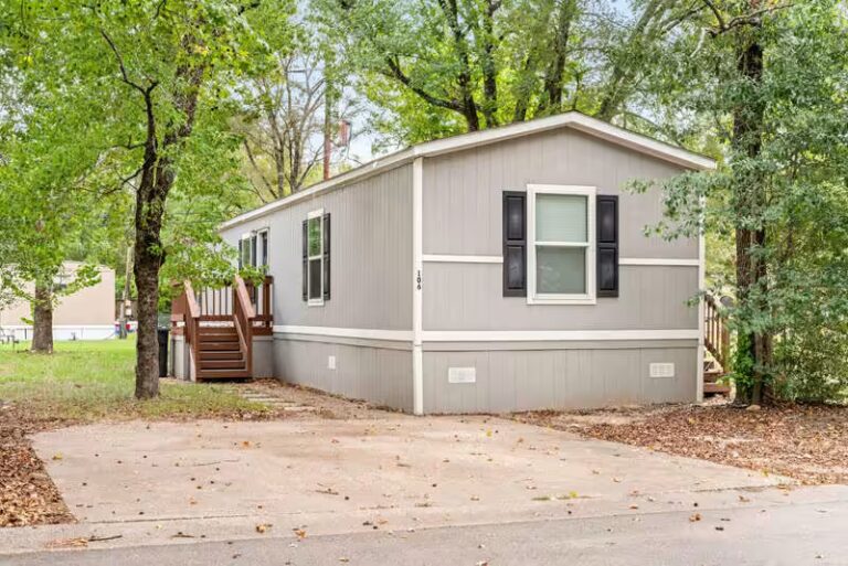 Gray mobile home marked 105 with black shutters, wooden deck stairs, and concrete driveway surrounded by trees