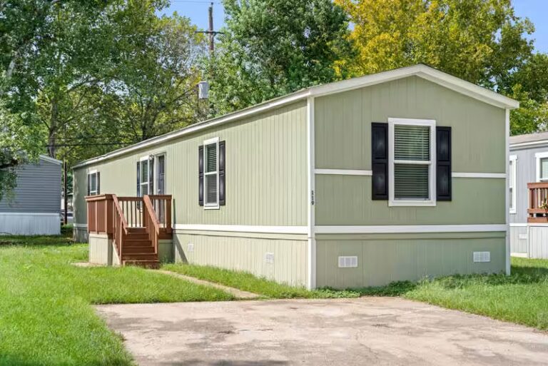 Green single-wide mobile home with black shutters, white trim, and wooden entry stairs on grassy lot.