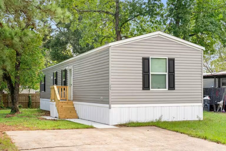 Gray single-wide mobile home with black shutters and wooden steps surrounded by trees.