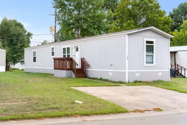 Gray single-wide mobile home with wooden deck stairs on grassy lot with concrete driveway pad