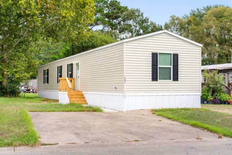 Single-wide mobile home with beige siding, black shutters, and wooden entry steps among trees.