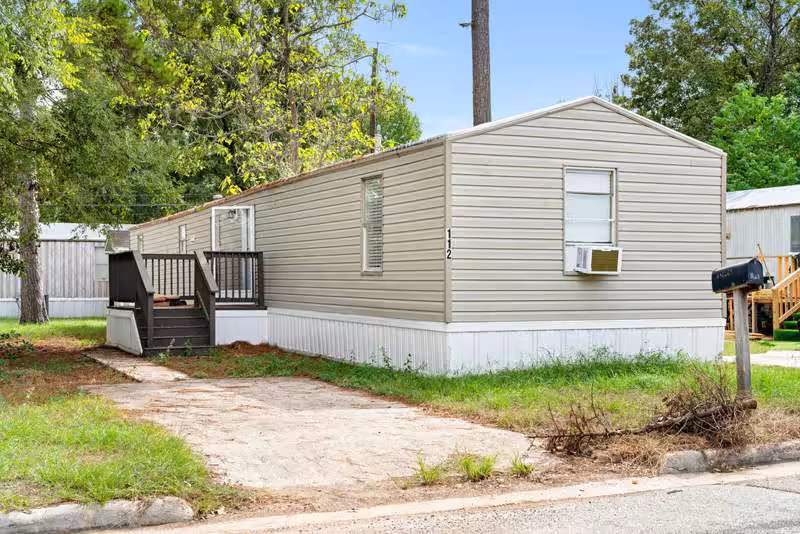 Beige mobile home marked 112 with wooden deck and stairs, window AC unit, surrounded by trees.