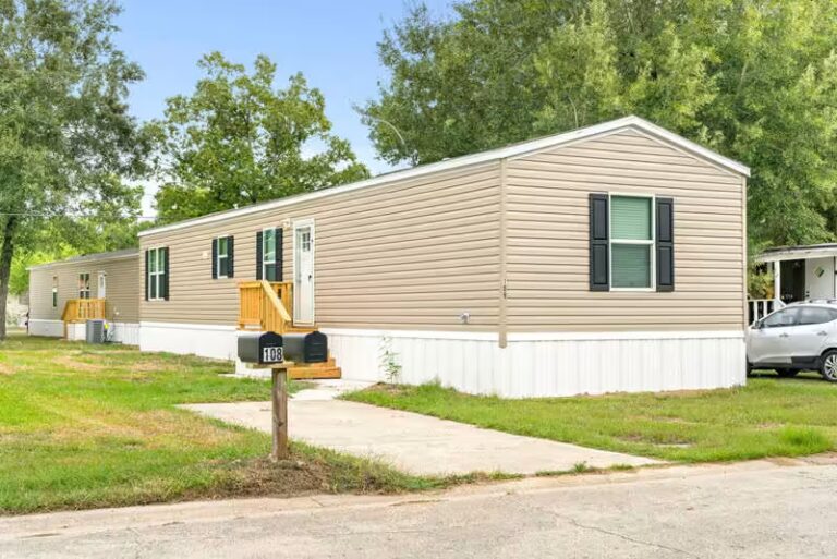 Beige mobile home with wooden ramp and mailbox numbered 108, surrounded by trees and grass.