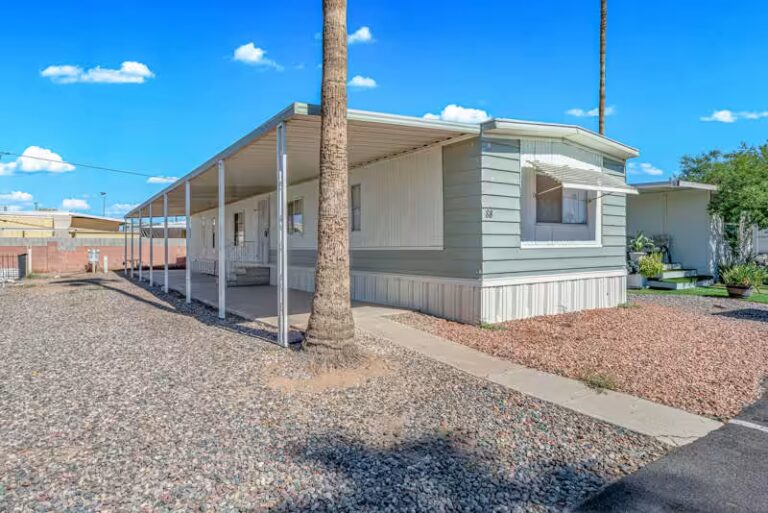 Mobile home with covered patio, palm tree trunk, and gravel yard under a blue sky.