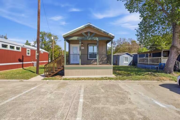 Small tan house with green-trimmed covered porch and wooden stairs beside a parking lot