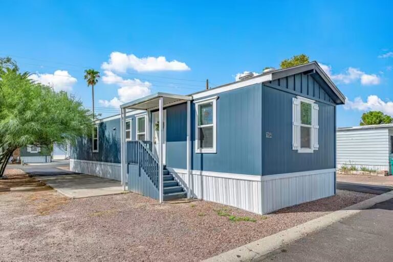 Blue mobile home with white trim, covered porch, and stairs, numbered 141, under a bright blue sky.