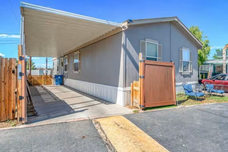 Gray mobile home with white carport awning, wooden fence gate, and blue patio chairs beside a driveway.