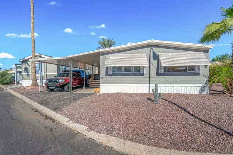 Gray mobile home with carport sheltering a red truck amid desert landscaping and palm trees.