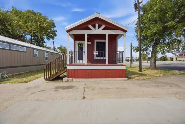Small red cabin with white-trimmed porch and wooden stairs beside a larger mobile home.