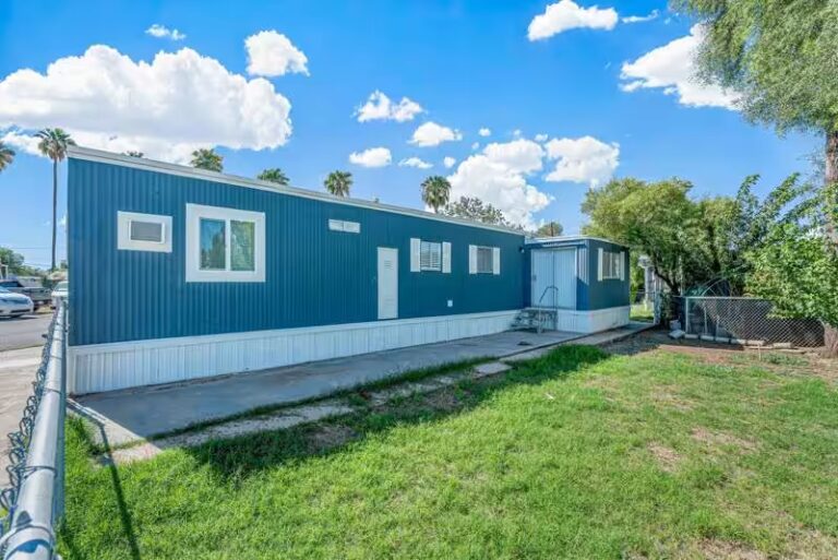 Blue mobile home with white trim under sunny sky with palm trees and green lawn.