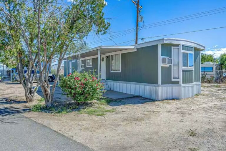 Green mobile home with white trim and carport, flowering bush, and tree on sandy lot under blue sky