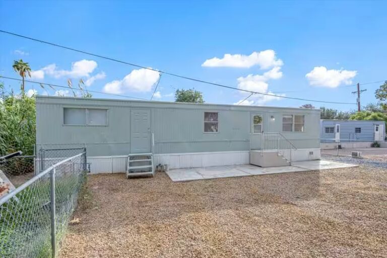 Pale green single-wide mobile home with gravel yard, chain-link fence, and blue sky with clouds.