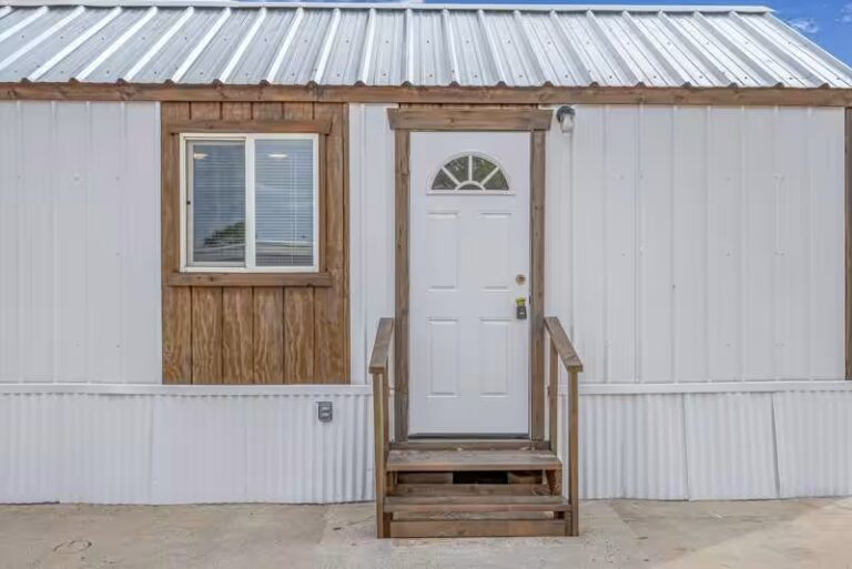 White metal building with wooden entry door, arched window, small side window, and wooden steps.